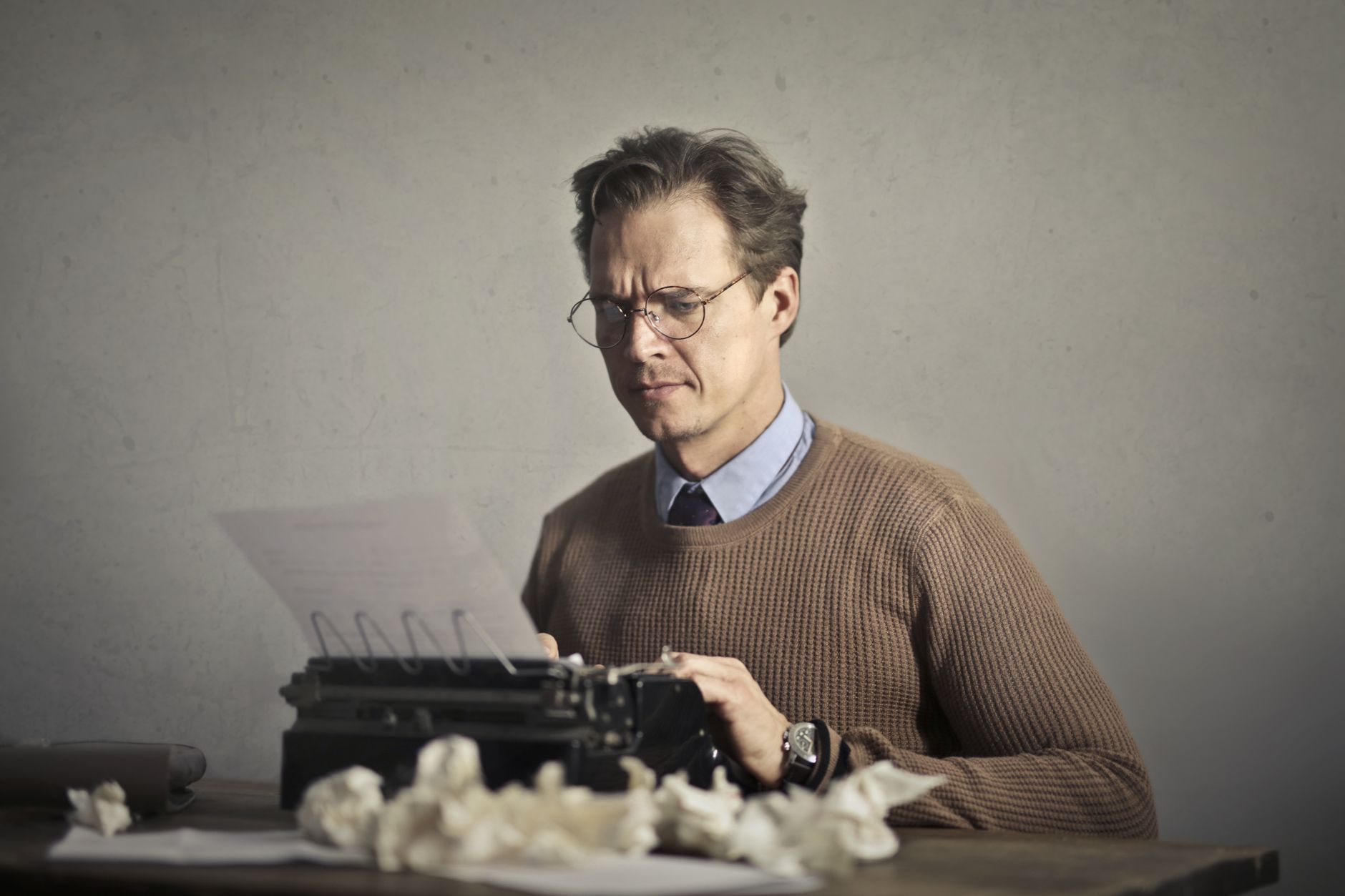 Pensive displeased man in glasses and elegant clothes sitting at table and using typewriter among mush of crumpled sheets of paper while exp