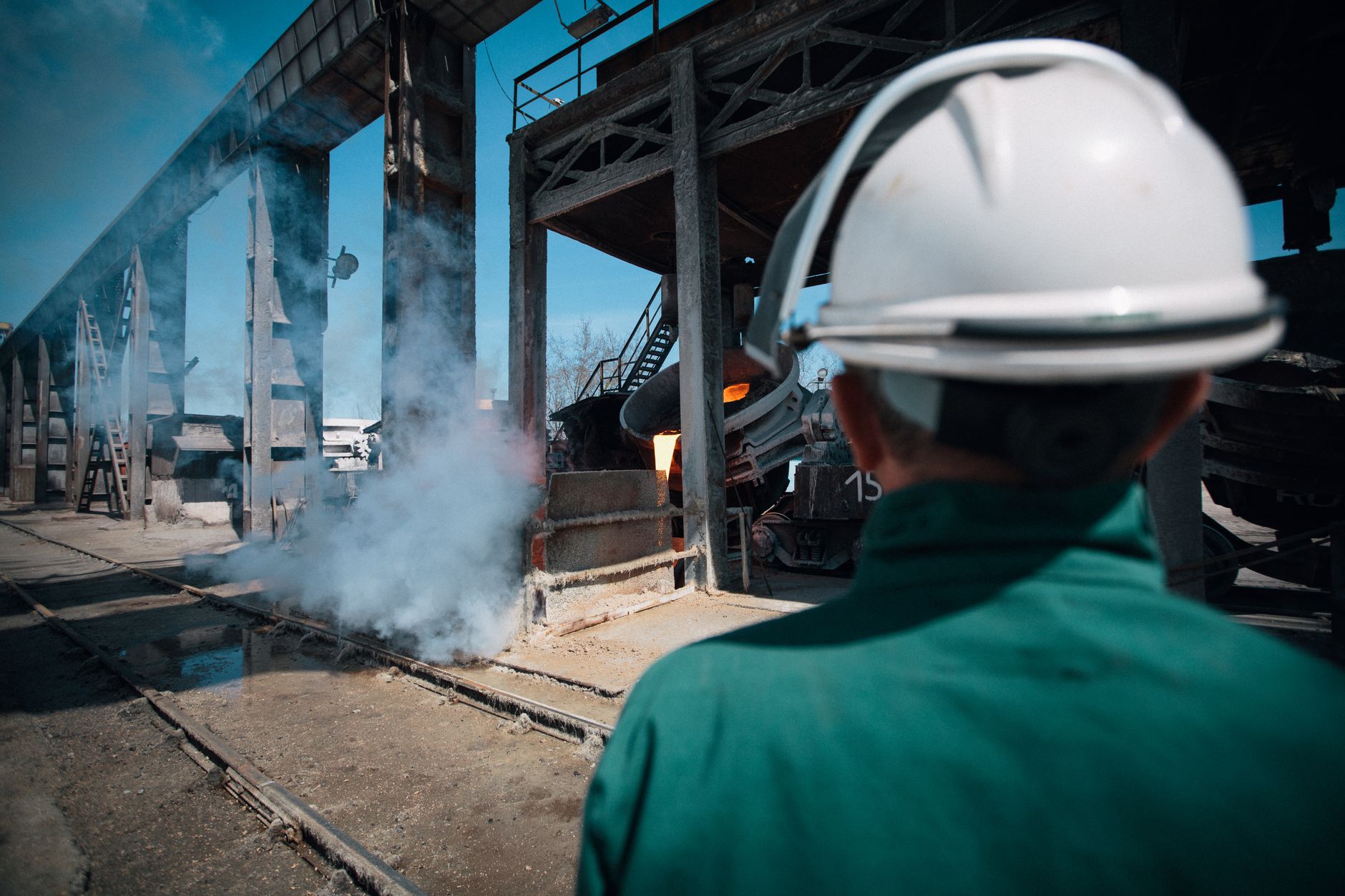 Factory worker in a hard hat watches molten metal pouring at an industrial site.
