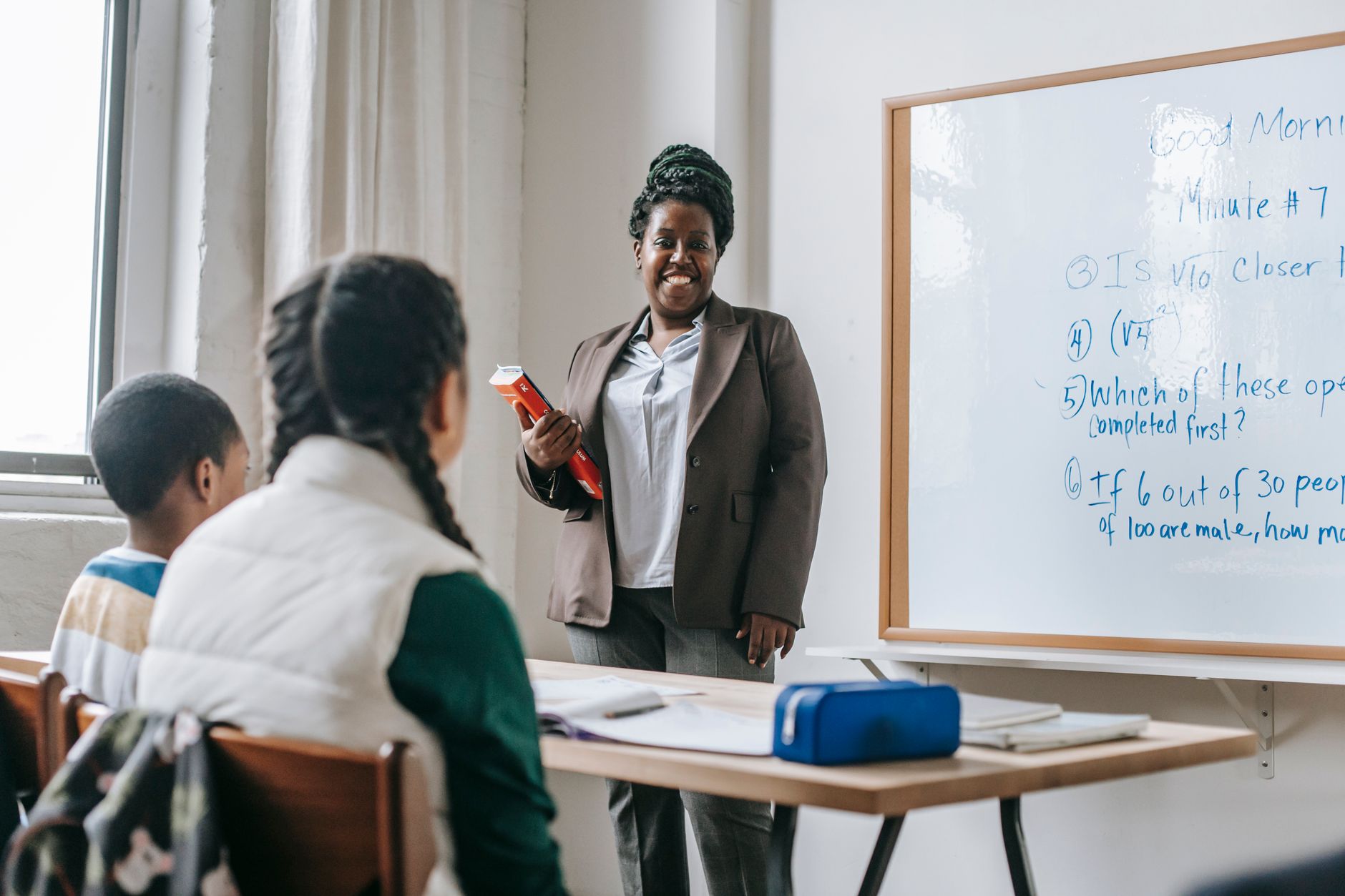Happy black female teacher standing near whiteboard and explaining lesson to unrecognizable children sitting at wooden table with books and 