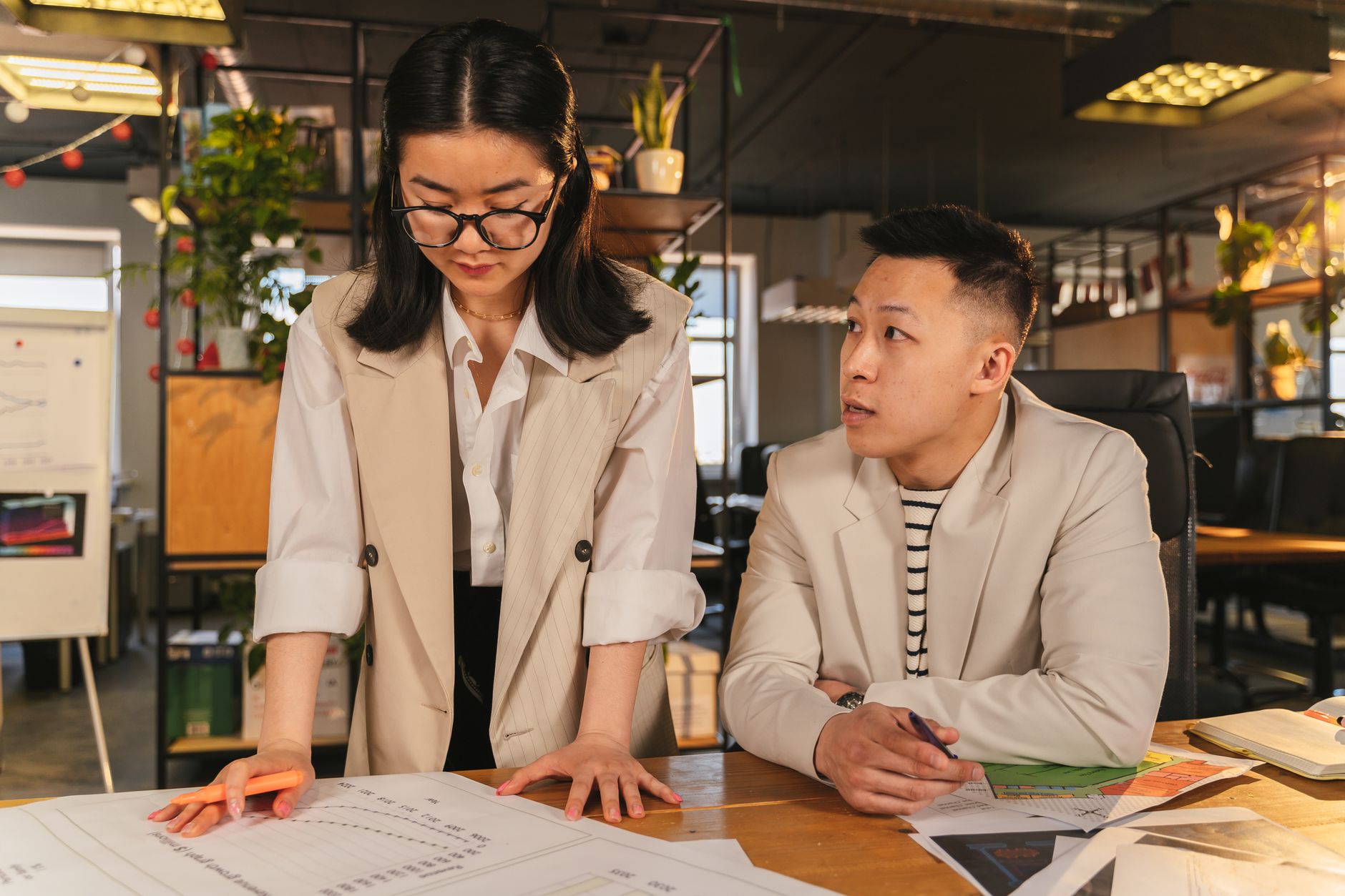Two colleagues engaged in a focused discussion in a modern office setting with plants and documents.