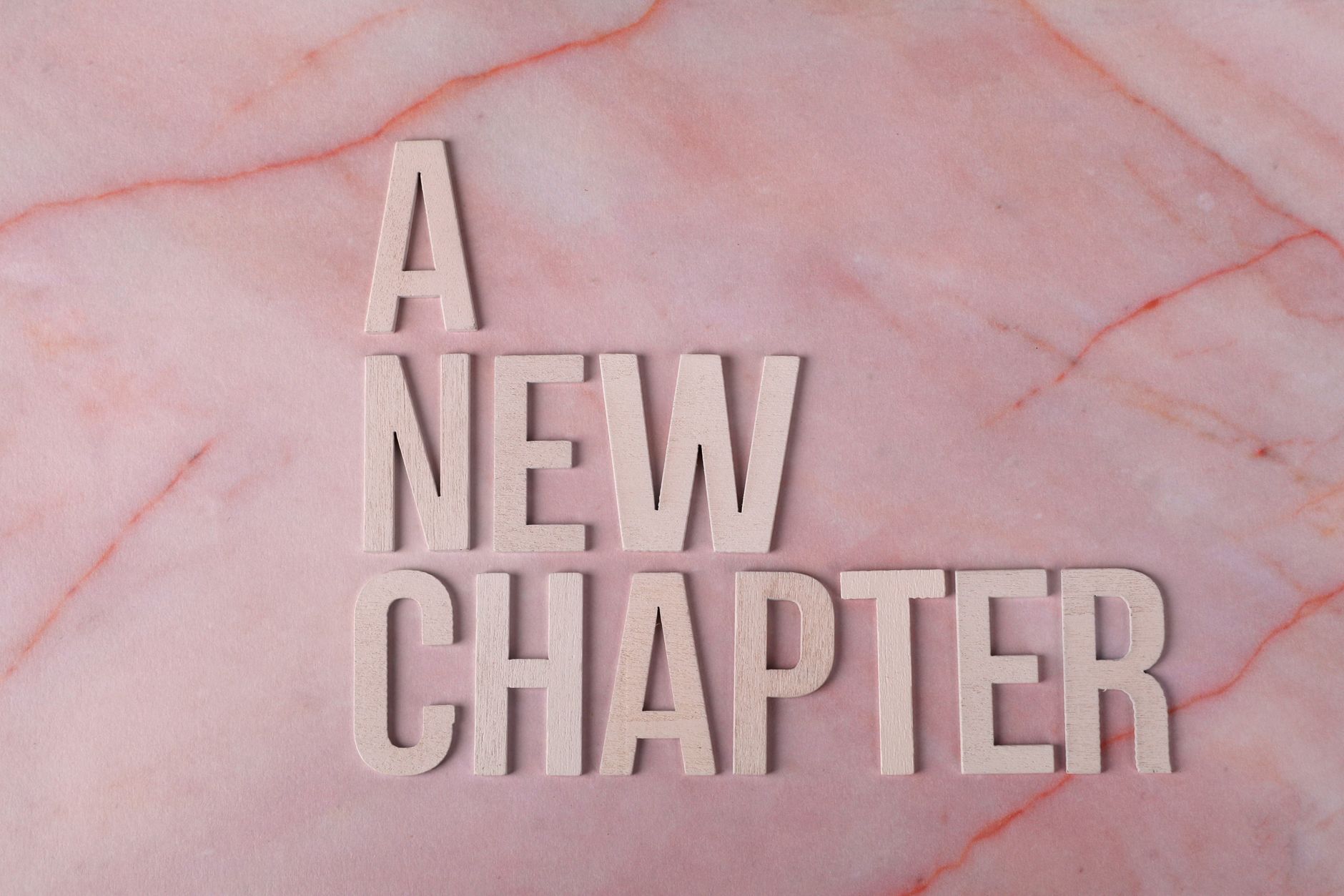 Wooden letters spell 'A New Chapter' on a pink marble background, symbolizing beginning.