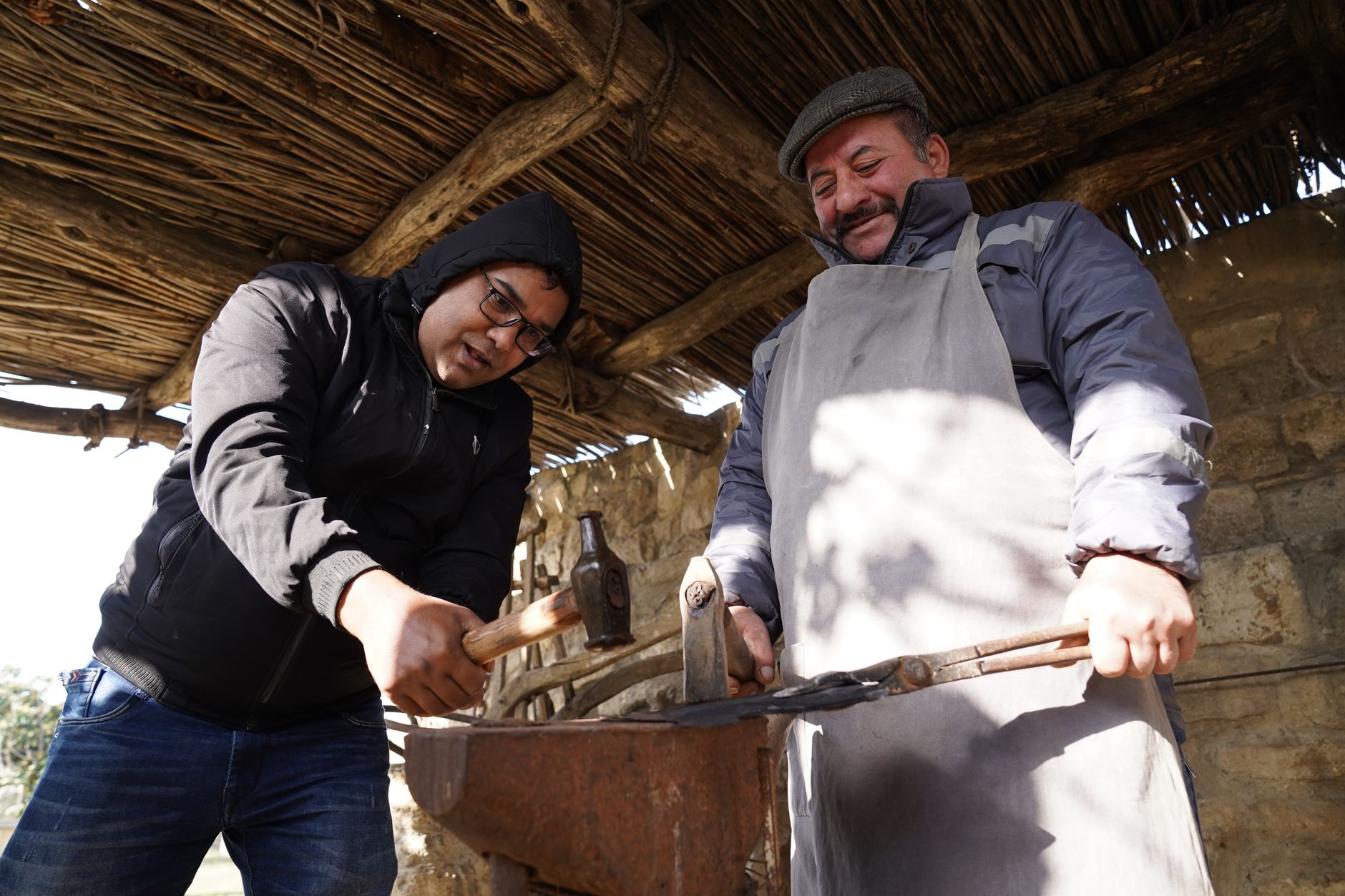 Two men collaborating in a blacksmith shop, working with hammers and an anvil under a rustic roof.