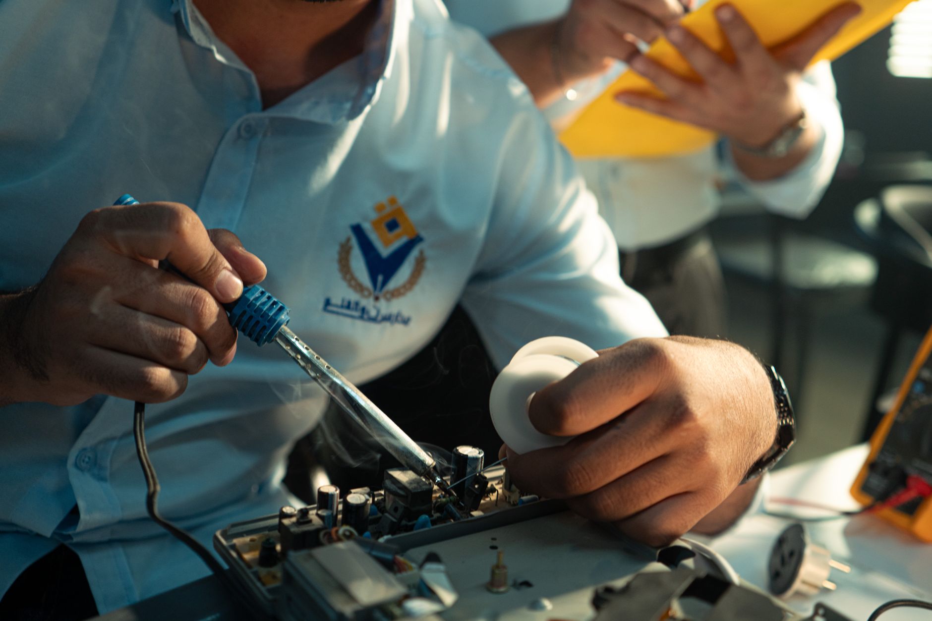 A technician engaged in soldering electronic components in Baghdad, Iraq, wearing a blue logo shirt.
