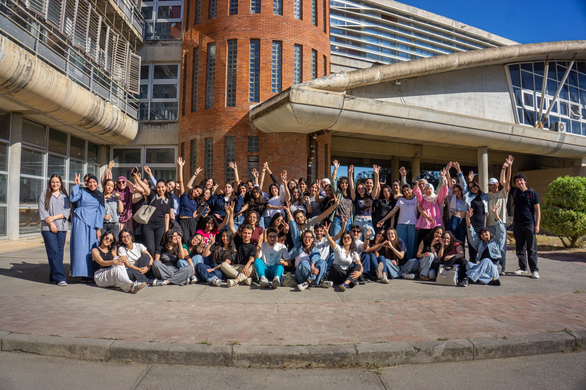 Large group of diverse young adults celebrating in front of a modern campus building on a sunny day.