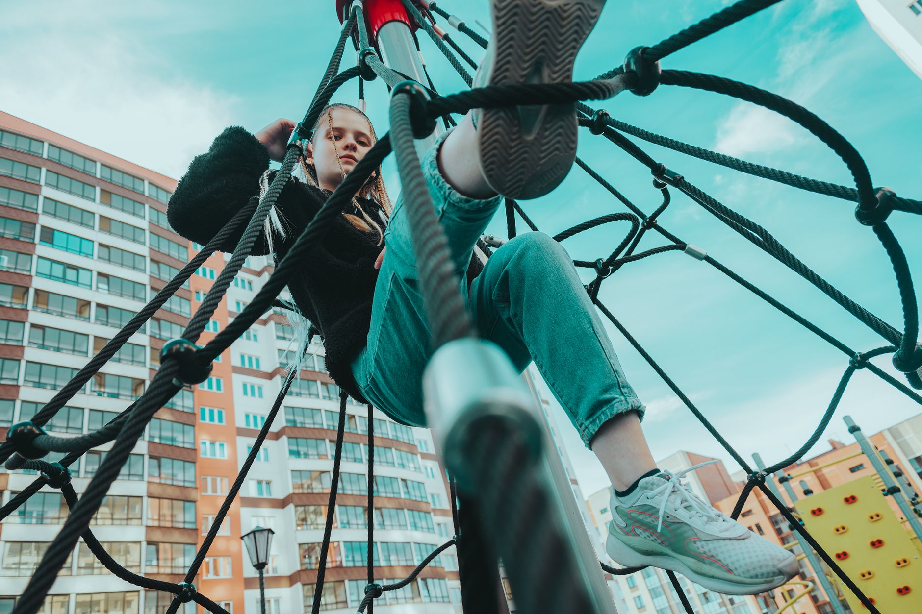 Low angle view of teenage girl climbing rope at playground with urban skyline backdrop.