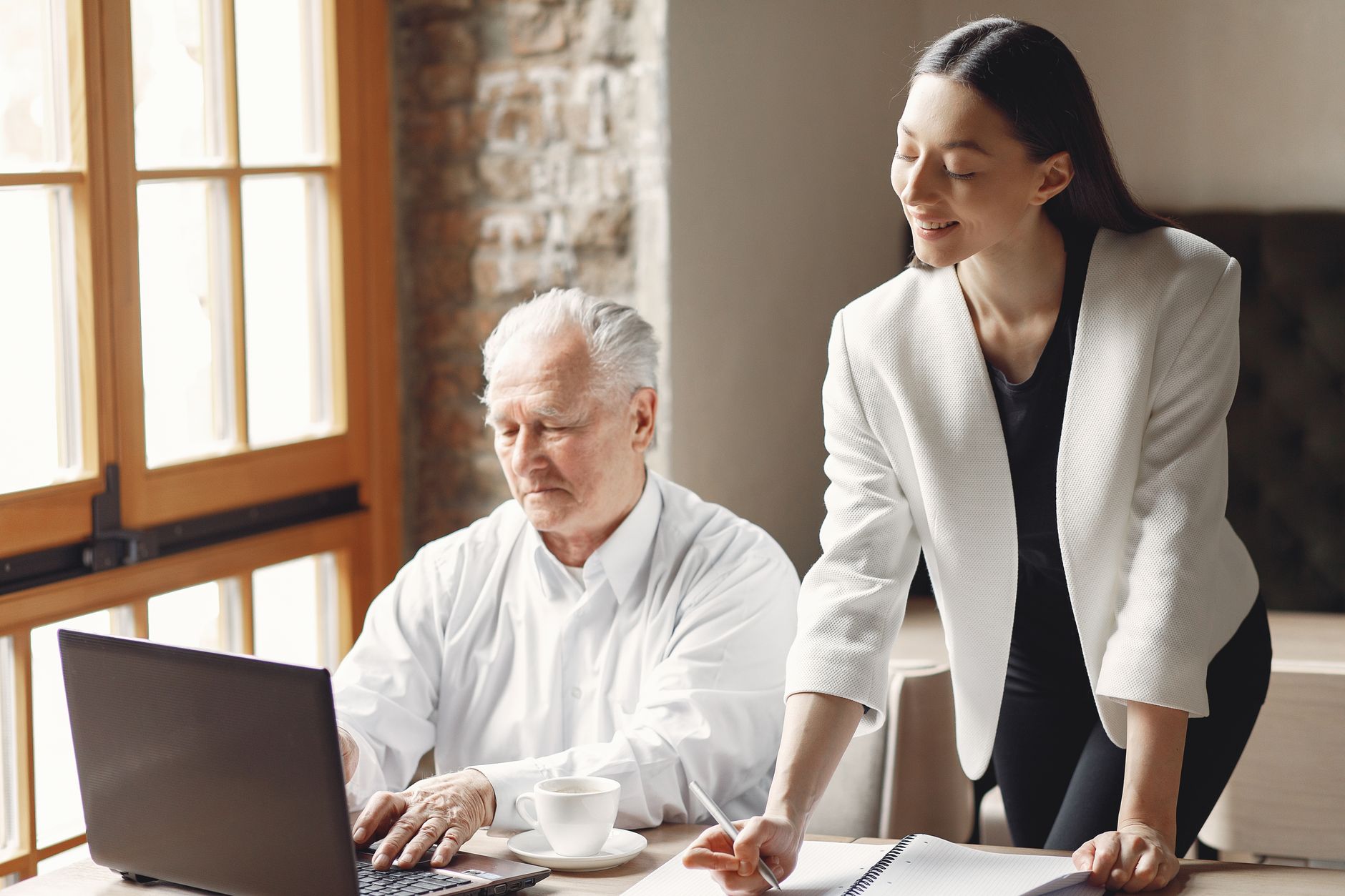Smiling young woman in formal wear standing and taking notes while stylish senior man working on laptop in light workspase at daytime