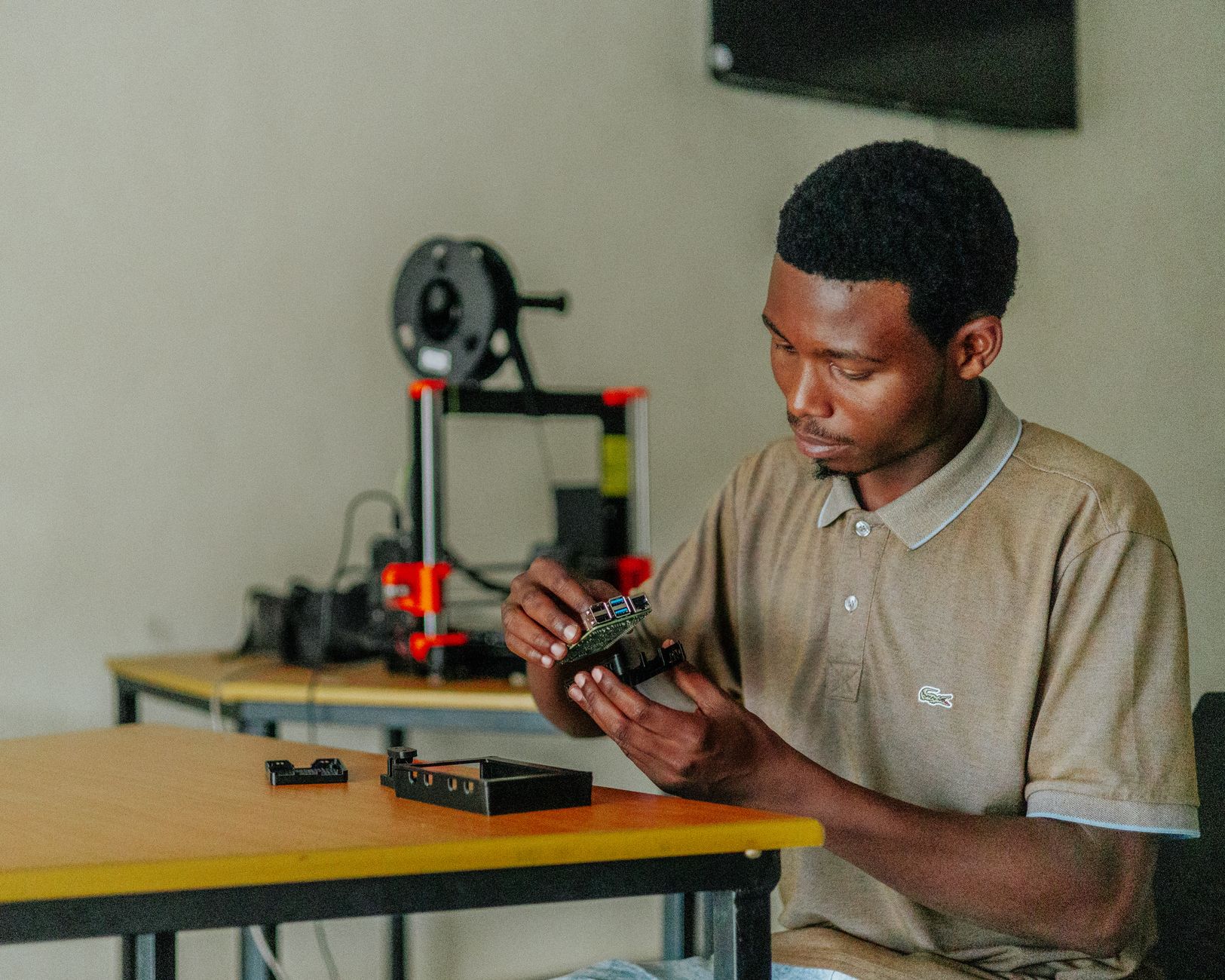 Focused young man assembling electronics with a 3D printer in the background.
