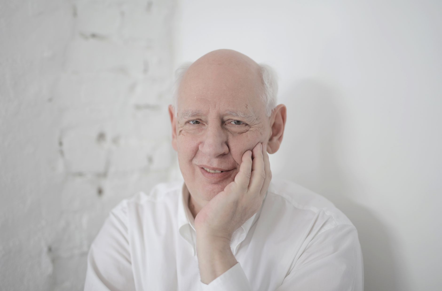 A thoughtful elderly man with white hair, resting his face on his hand, dressed in a white shirt.