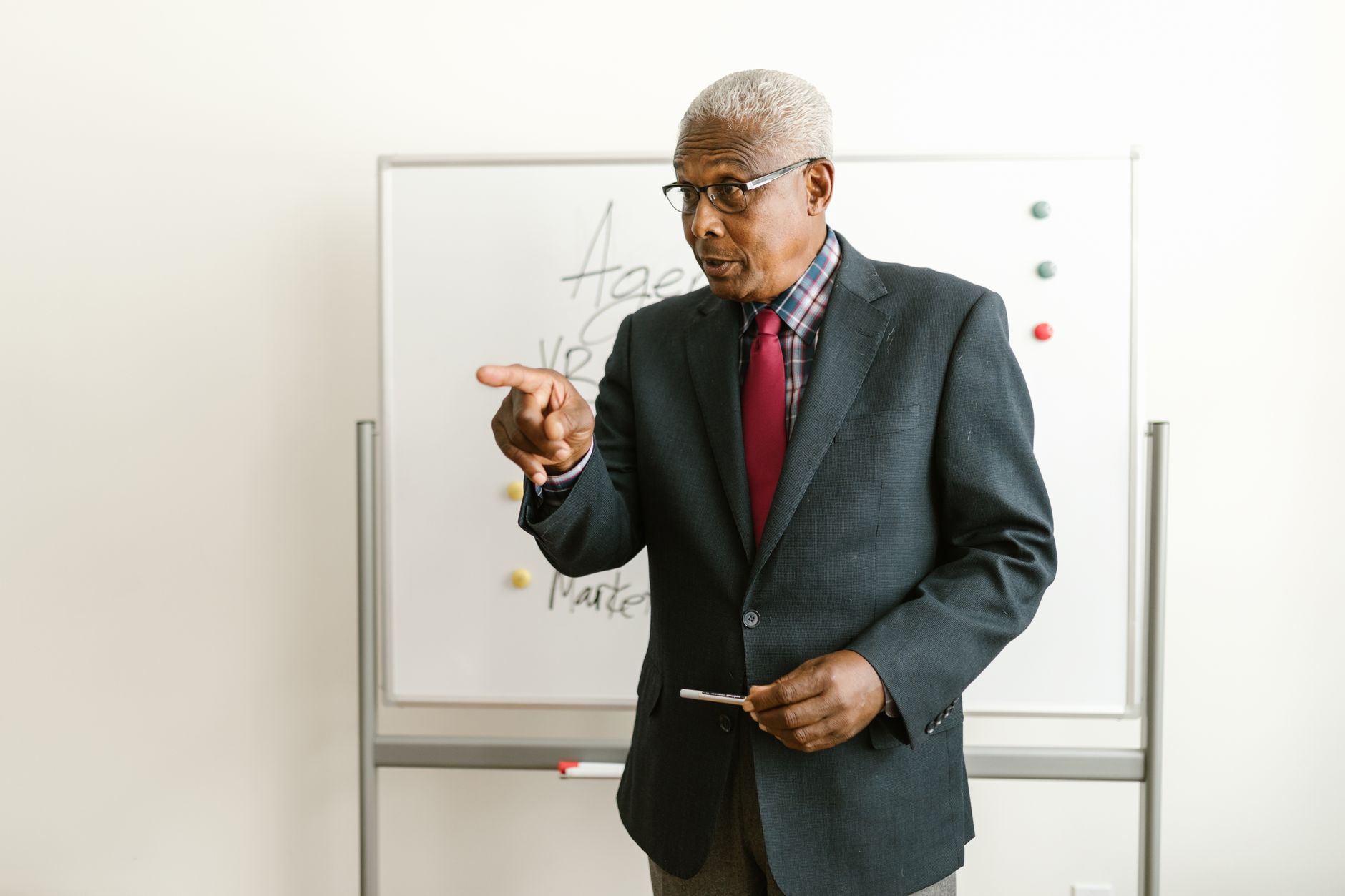 Elderly man in business attire giving a presentation in front of a whiteboard.