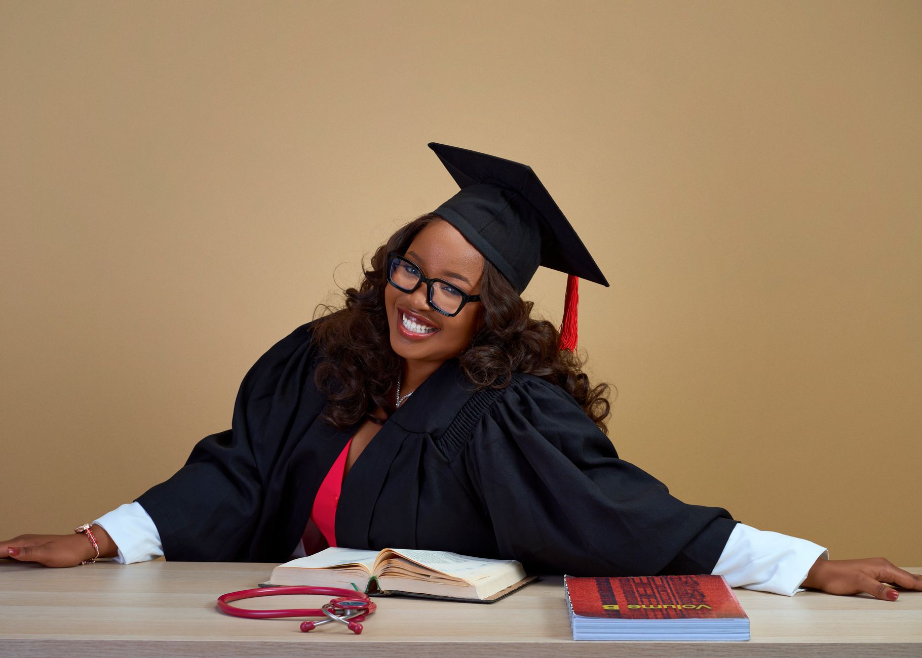 Happy graduate in cap and gown smiling with books and stethoscope on desk.
