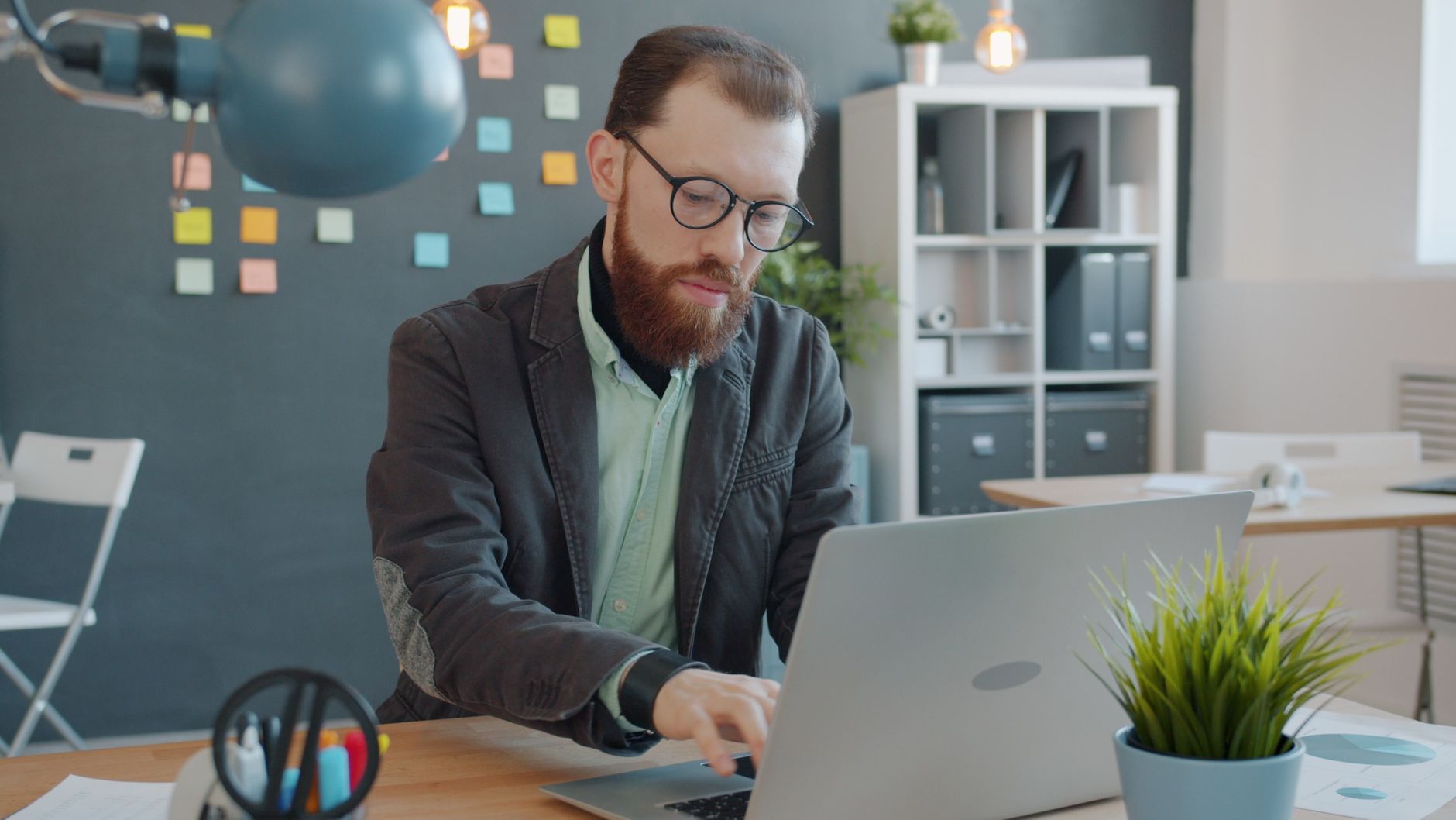 Man typing on a laptop in a modern office environment with sticky notes in the background.