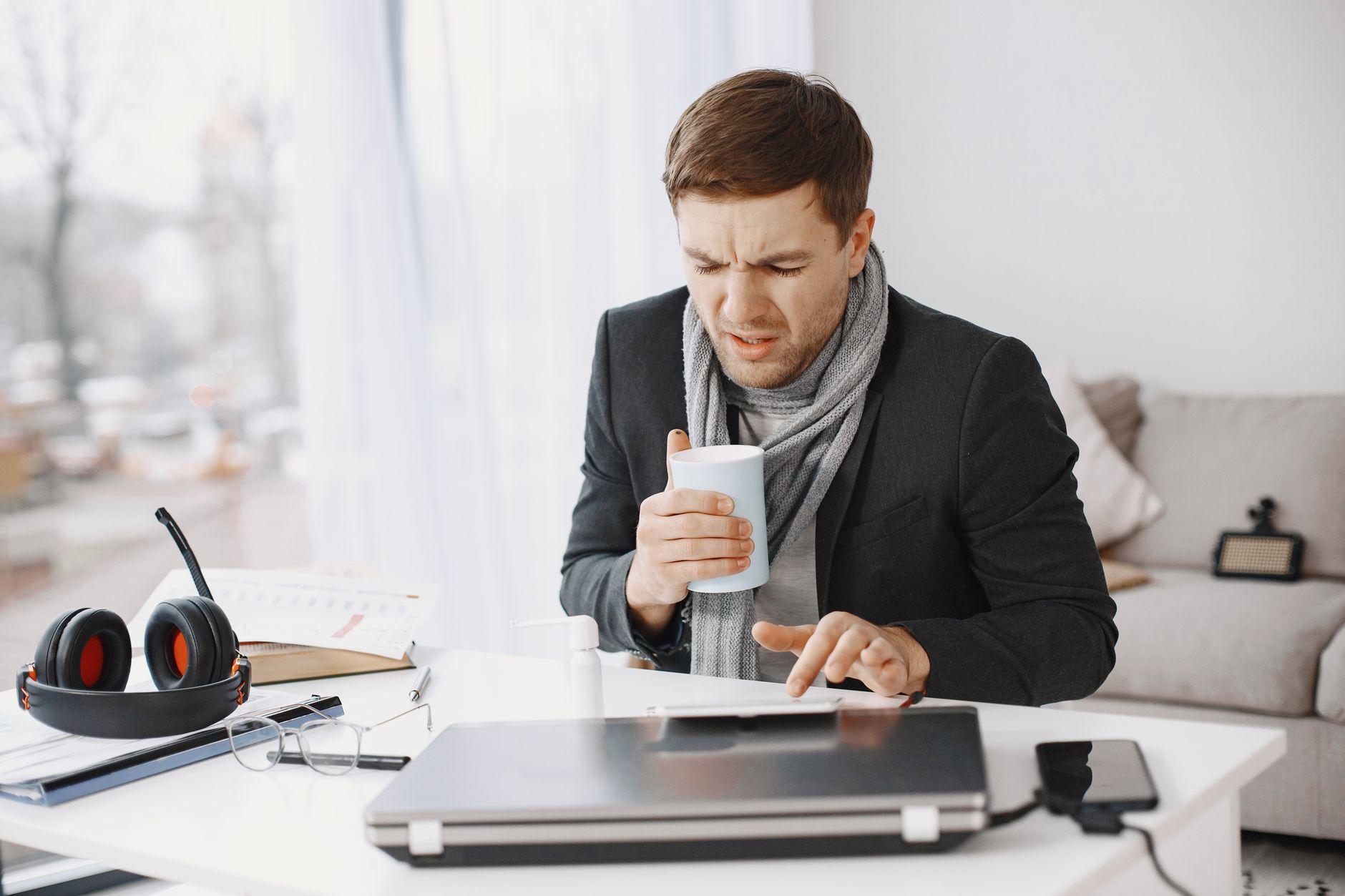Unwell businessman working at home holding tea while sitting at a desk with a laptop.