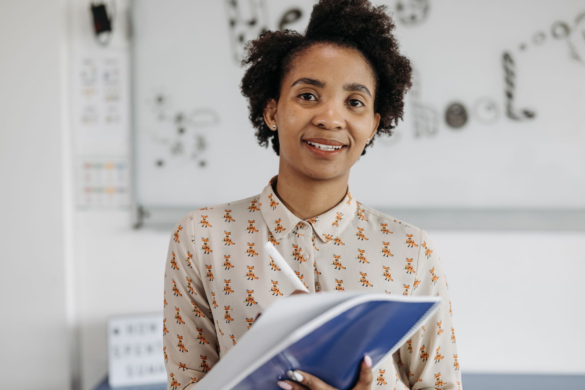 Confident female teacher smiling while holding a notebook in a classroom setting.